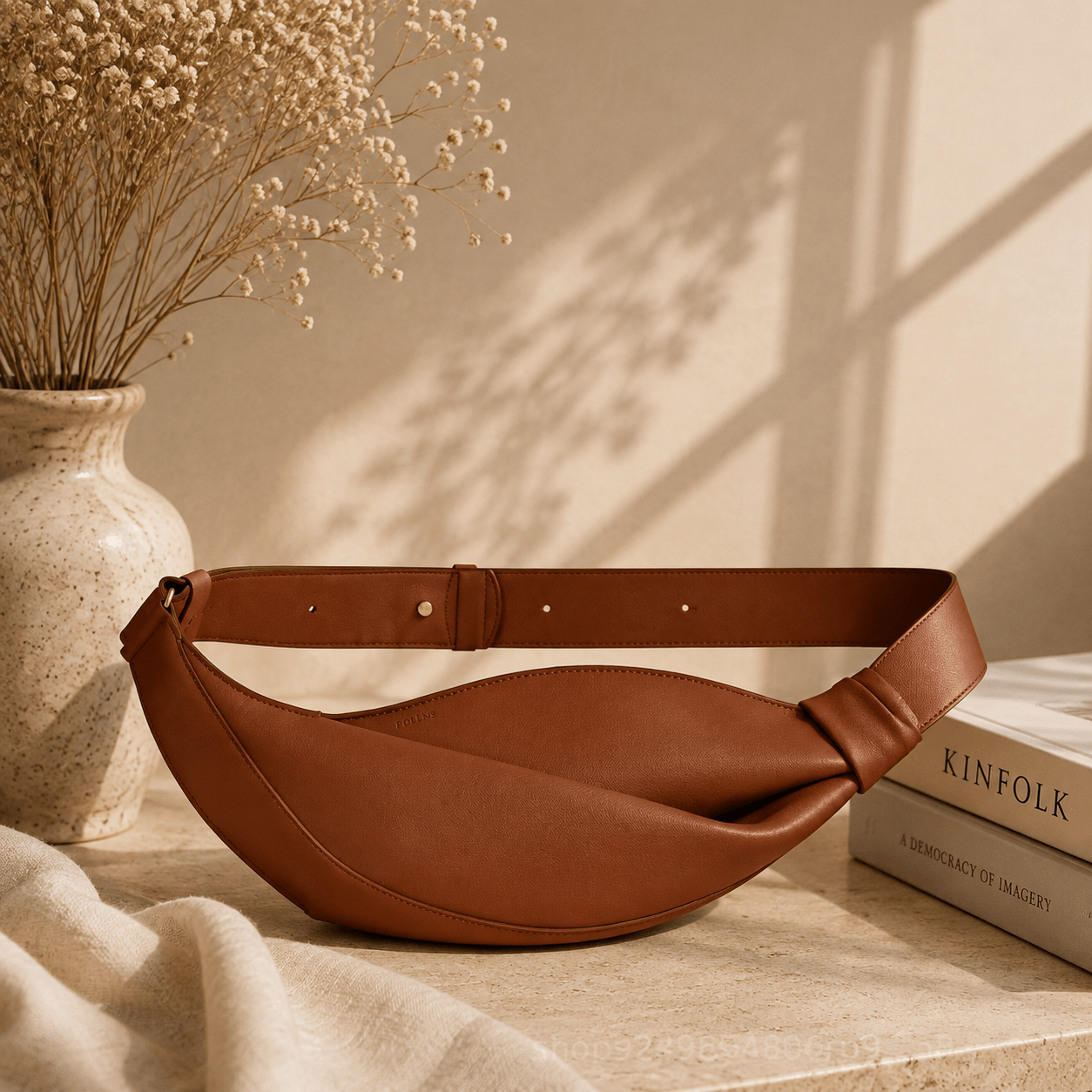 A brown leather crescent-shaped belt bag with a matching adjustable strap, styled on a stone surface next to a vase of dried flowers and a book.