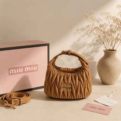 A Miu Miu brown quilted leather handbag with a top handle and gold logo, displayed next to its pink box, matching strap, and tags on a beige surface with dried flowers in a vase.