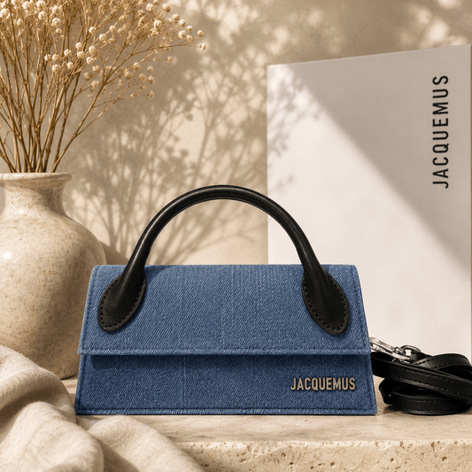 A blue denim handbag with black leather handles and a black shoulder strap, styled on a stone surface with a beige vase of dried flowers and a white paper bag in the background. The brand name 'JACQUEMUS' is visible on the bag and the paper bag.