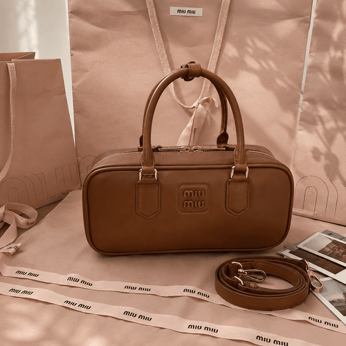A brown Miu Miu leather handbag with top handles and a matching detachable shoulder strap, styled on beige Miu Miu branded paper and fabric with branded ribbon. The Miu Miu logo is embossed on the front of the bag.