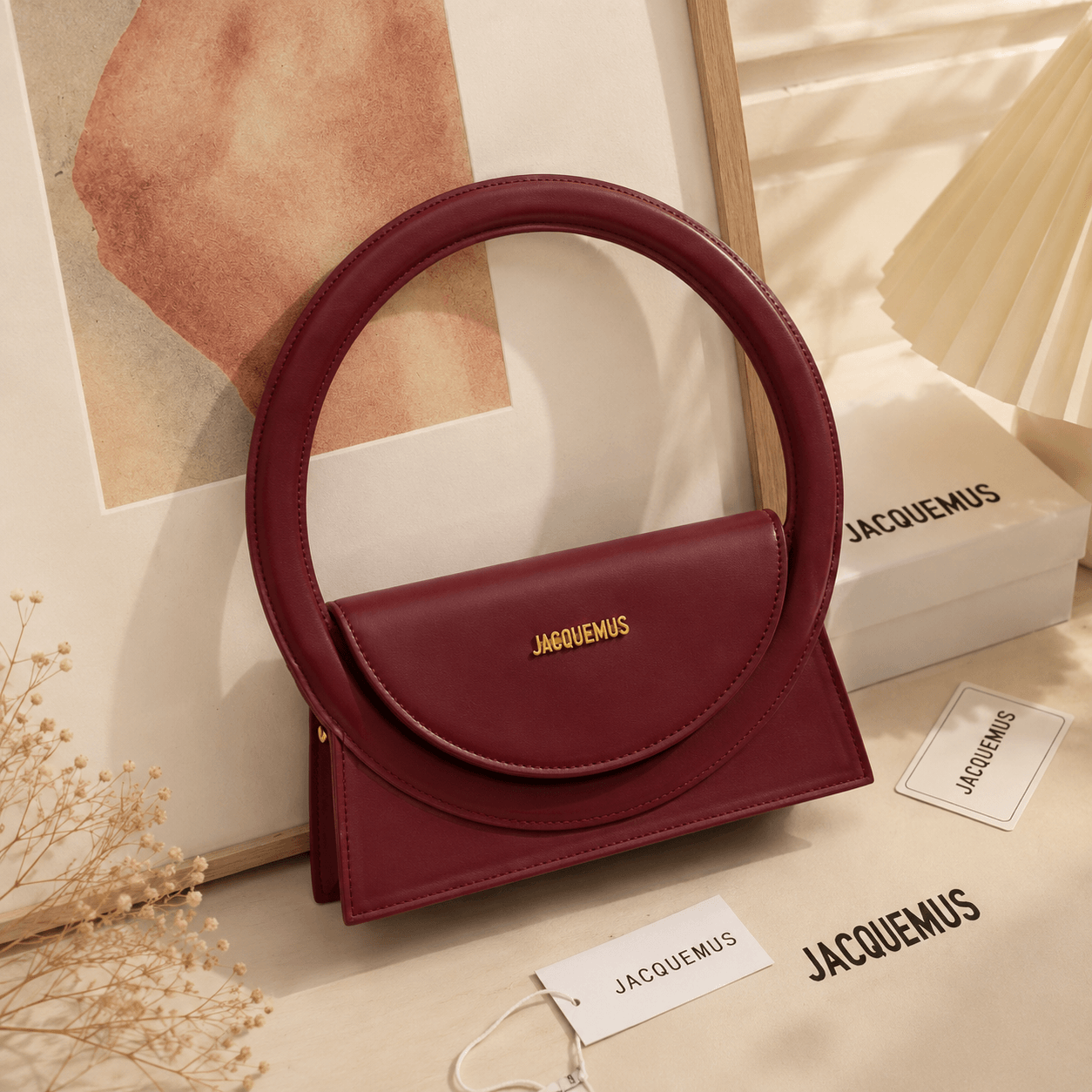 A burgundy leather handbag with a circular top handle and a flap closure featuring a gold jacquemus logo, styled on a beige surface with branded tags, a box, and a framed print.