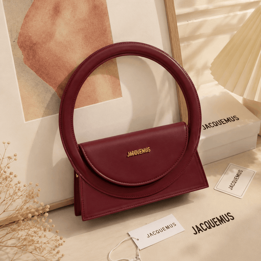 A burgundy leather handbag with a circular top handle and a flap closure featuring a gold jacquemus logo, styled on a beige surface with branded tags, a box, and a framed print.