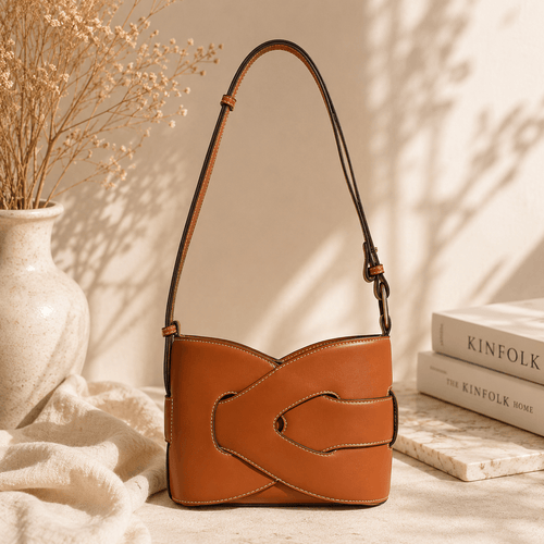 Classic cognac brown shoulder bag made from smooth faux leather featuring a distinctive wraparound buckle design, contrast stitching, adjustable shoulder strap, and structured silhouette displayed in a warm minimalist lifestyle setting.