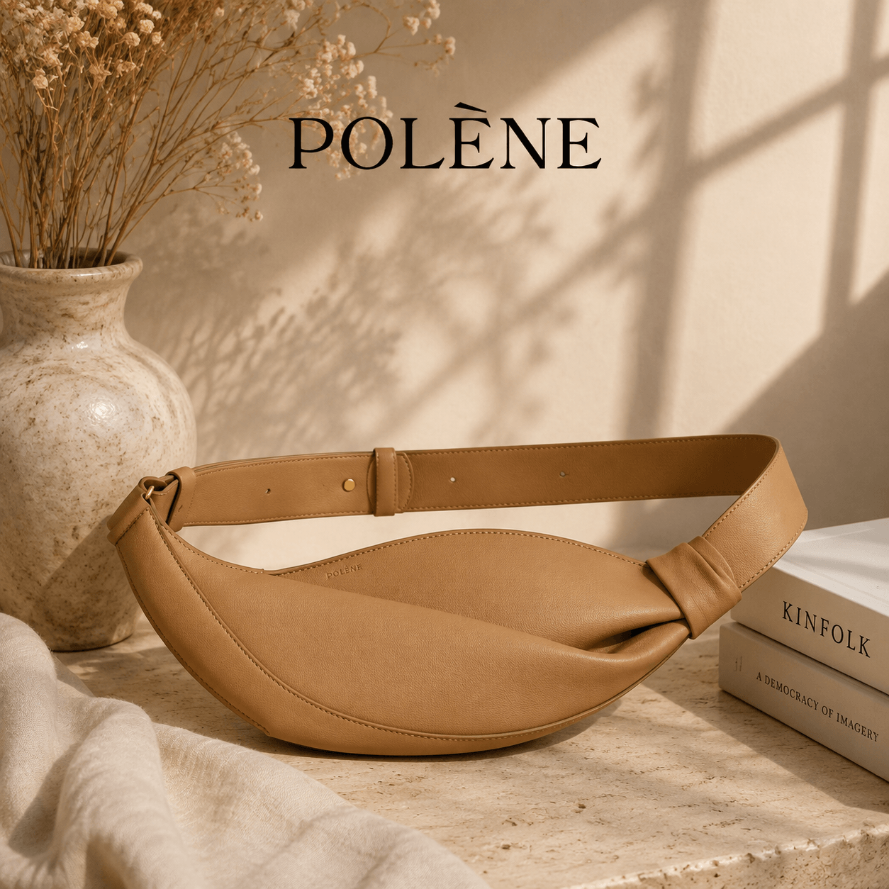 Minimalist camel leather crescent belt bag with a curved silhouette, adjustable wide strap, and elegant neutral design displayed in a warm beige aesthetic setting with natural sunlight and dried flowers.