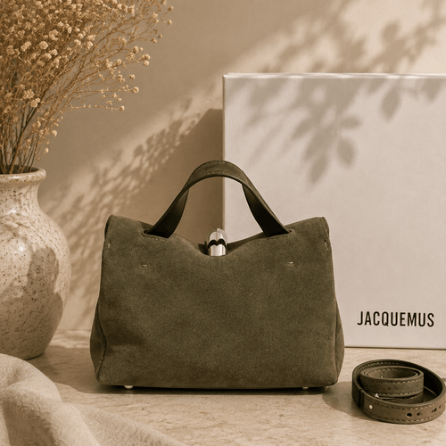 An olive green canvas handbag with dark green leather handles, displayed on a beige surface next to a white Jacquemus box, a vase with dried flowers, and a matching green belt.